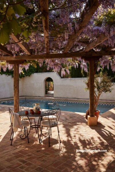 Vine covered pergola over terracotta patio beside spanish style pool with wrought iron furniture
