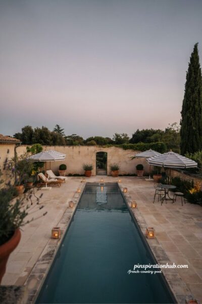 French country pool at dusk with striped umbrellas and lantern lighting by the water