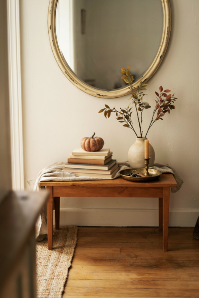 **Prompt:** **1. Shot type:** A professional editorial interior photography shot from a natural eye-level angle slightly across the entryway, framing the full console table and surrounding wall while placing the woven rattan tray toward one side of the tabletop as a clearly defined styling vignette. Use a vertical, mobile-friendly composition with enough negative space to show the balance between the low tray arrangement and a taller centerpiece. **2. Room description:** A bright, airy fall entryway with a warm natural wood console table beneath a simple mirror or understated artwork. A shallow woven rattan tray sits intentionally toward one side of the console, containing a small candle, petite ceramic vase, and a few scattered acorns. The opposite side holds a taller arrangement of dried autumn branches, creating a relaxed height contrast while keeping the tabletop open, functional, and visually balanced. **3. Style and era:** Warm organic farmhouse with relaxed coastal-meets-country character, combining natural woven textures, vintage-inspired accents, and understated contemporary fall styling. **4. Color palette:** Natural honey rattan, warm oak, oatmeal, creamy ivory, muted rust, soft caramel, dusty olive, earthy brown, and aged beige, creating a gentle autumn palette with plenty of natural warmth. **5. Key props and details:** A handmade-looking woven rattan tray with irregular natural fibers and softly rounded edges, containing only two or three carefully chosen objects: a small cream pillar candle, a petite weathered ceramic vase with a few dried stems, and several natural acorns scattered loosely inside the tray. Add a taller stoneware vase filled with dried autumn branches toward the opposite side of the console, along with a softly textured linen runner, a small vintage bowl for keys, and subtle everyday objects. Keep every vignette element contained within the tray's edges and include natural imperfections such as uneven rattan fibers, slightly weathered ceramic, gently wrinkled linen, visible wood grain, and casually arranged acorns. **6. Lighting:** Soft, warm natural sunlight filtering through a nearby window or doorway, casting delicate shadows from the rattan weave and dried branches across the console surface. A small candle adds a subtle amber glow without overpowering the daylight, creating the feeling of a peaceful autumn afternoon. **7. Mood and atmosphere:** Warm, organized, earthy, and effortlessly inviting. The soft autumn colors should immediately make the entryway feel calmer, while the rough woven rattan, worn wood, handmade ceramic, dried botanicals, and linen add warmth and depth against harder surfaces. The contained tray vignette should feel intentional without looking overly styled, showing how a simple boundary can turn a few everyday objects into a polished seasonal arrangement. The overall scene should communicate easy, practical fall decorating that feels collected, flexible, and genuinely lived-in. **8. Technical tail:** no people, no text, no watermarks, no blur, no bokeh, all surfaces in sharp focus, photorealistic, 4K resolution, editorial interior photography, Pinterest-worthy